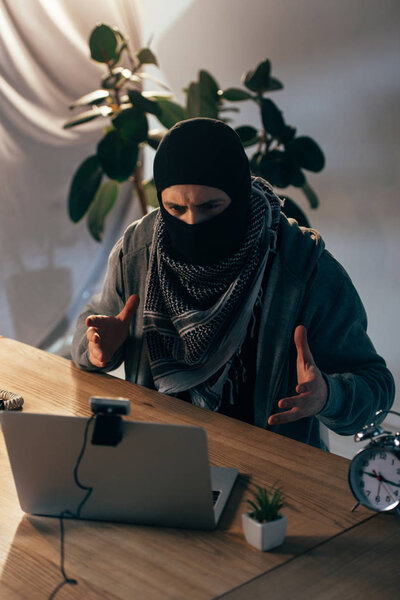 Злой преступник в маске жестом на веб-камеру в комнате
