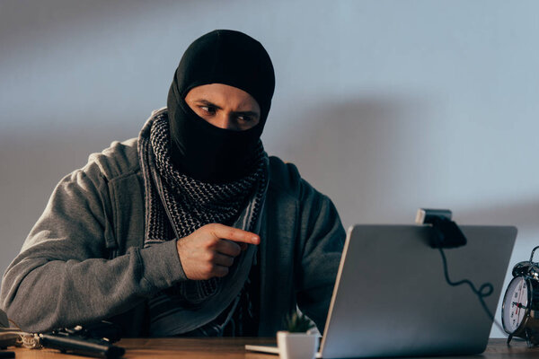 Террорист в маске смотрит на веб-камеру и показывает пальцем на экран ноутбука
