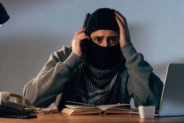 Worried terrorist in mask with book and gun touching head
