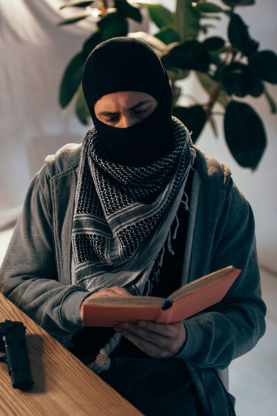 Serious terrorist in black mask reading book at table