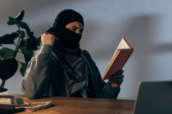 Serious terrorist in mask with gun reading book in room