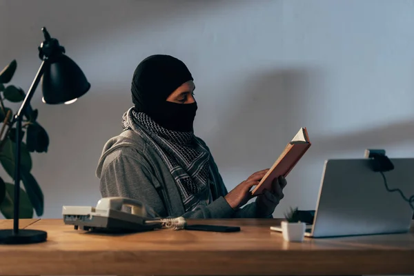 Konzentrierter Terrorist Mit Schwarzer Maske Sitzt Tisch Und Liest Buch — Stockfoto