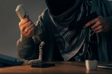 Cropped view of terrorist holding handset and gun at table clipart