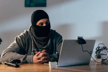 Terrorist in mask sitting with interlaced fingers and looking at webcam clipart