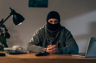 Terrorist in mask sitting at table with interlaced fingers clipart