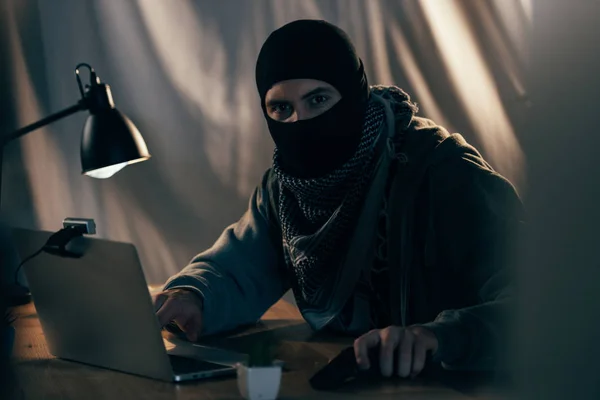 恐怖分子戴着面具 带着枪在黑暗的房间里使用笔记本电脑 — 图库照片