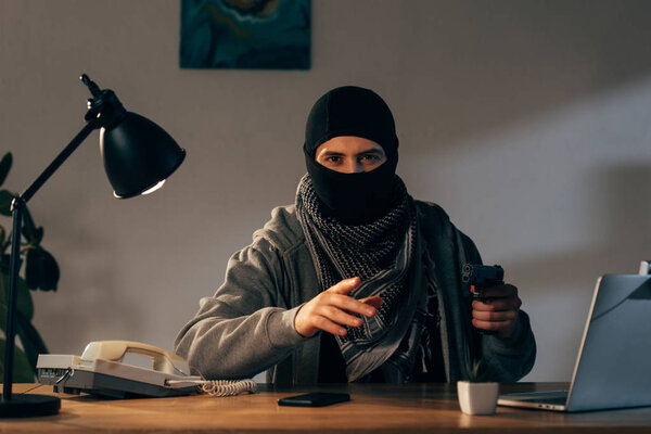 Terrorist in mask sitting at table with lamp and holding gun