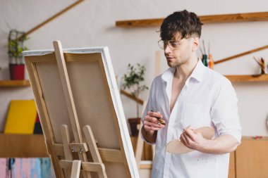 handsome artist in glasses standing at easel with canvas in painting studio clipart