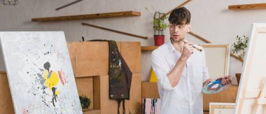 panoramic shot of handsome artist with paintbrush and palette looking at painting in painting studio clipart