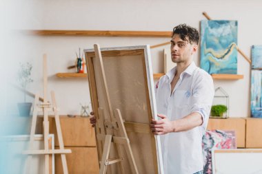 selective focus of handsome artist looking at camera while standing at easel with canvas  clipart