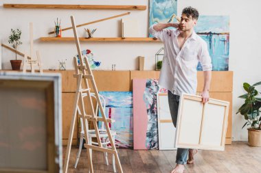 selective focus of handsome barefoot artist carrying canvas in painting studio clipart