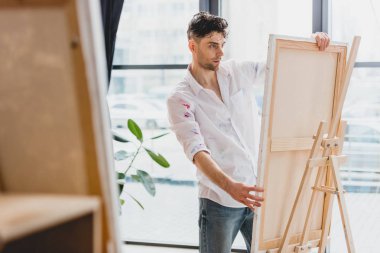 selective focus of handsome artist fixing canvas on easel in gallery clipart