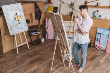 handsome artist in white shirt and blue jeans standing at easel in painting studio clipart