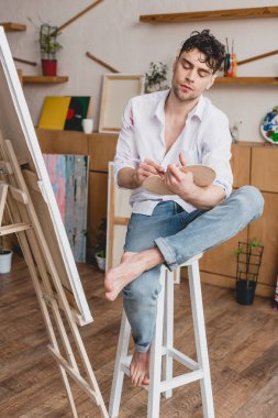 beyaz gömlek ve mavi jeans şövale resim Studio oturan yakışıklı sanatçı