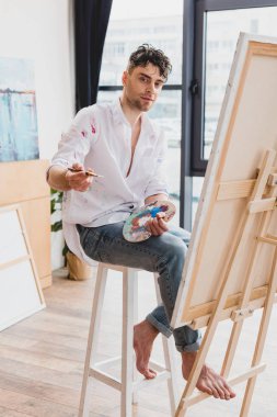 Palet ve yüksek sandalyede oturan ve kameraya bakarak boya fırçası ile yakışıklı sanatçı