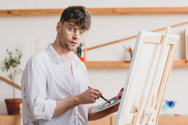 красивый художник в очках, стоящий у мольберта с палитрой и кистью и смотрящий в камеру
