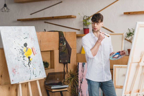 handsome artist with paintbrush and palette looking at painting in gallery