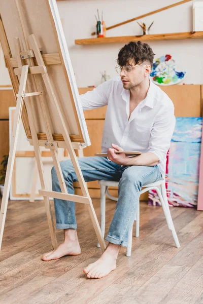 handsome artist in white shirt and glasses painting on canvas while sitting on chair in painting studio