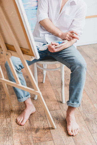 partial view of barefoot artist sitting at easel with canvas