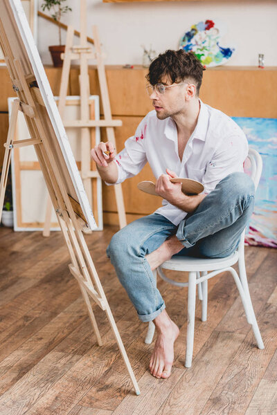 concentrated handsome artist painting on canvas while sitting on chair in gallery