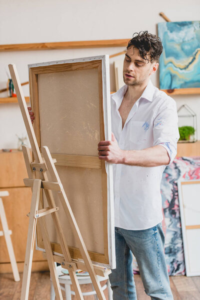 handsome artist in white shirt fixing canvas on easel in painting studio