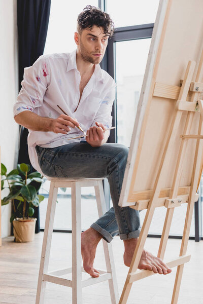 handsome artist in white shirt and blue jeans mixing paints on palette while sitting at easel in gallery