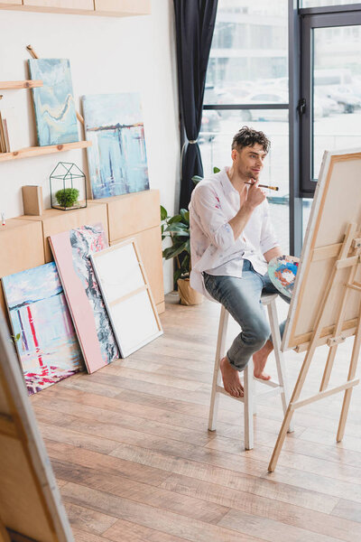 selective focus of smiling artist sitting on high chair at easel with canvas
