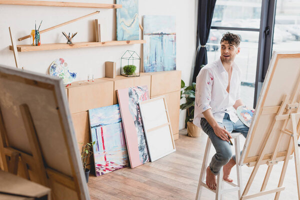 selective focus of handsome smiling artist sitting on high chair at easel 
