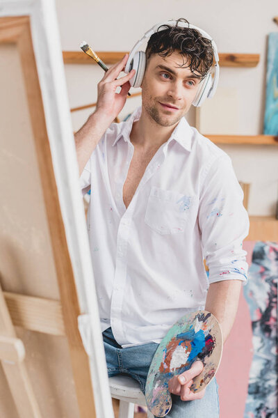 selective focus of handsome artist in headphones listening music while sitting at easel with canvas