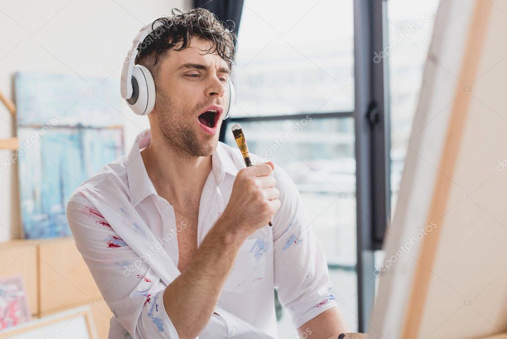 selective focus of handsome artist in headphones, singing with paintbrush