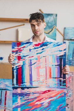 handsome artist holding multicolored abstract painting and looking at camera clipart