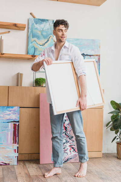 good-looking artist in white shirt and blue jeans carrying canvas in painting studio