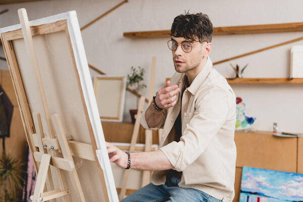 handsome artist in eyeglasses sitting near easel with canvas and looking at camera