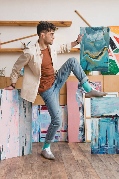 good-looking artist holding multicolored abstract paintings in painting studio
