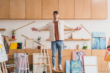 handsome artist standing on chair in spacious light painting studio clipart