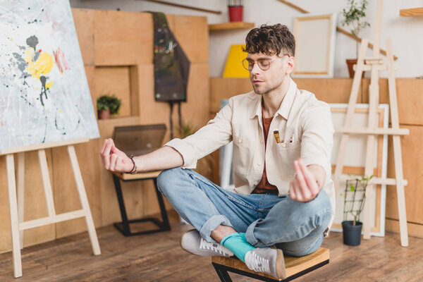 selective focus of dreamy artist with closed eyes sitting in lotus pose in gallery