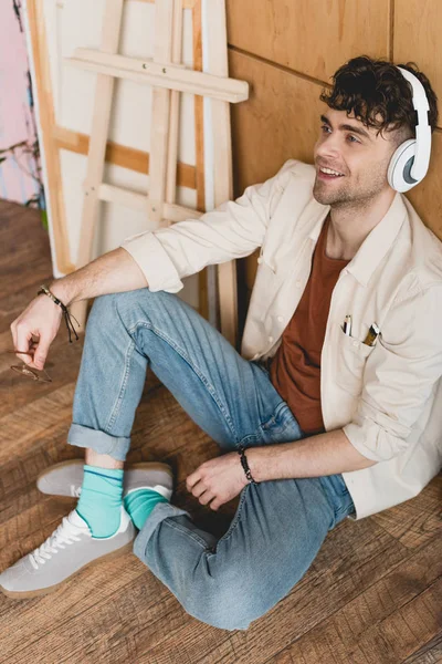 smiling artist in headphones sitting on floor and listening music in painting studio