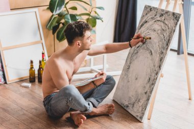 handsome half-naked artist sitting with crossed legs on floor and painting picture on canvas clipart