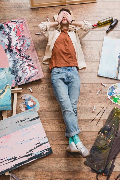 overhead view of tired artist lying on wooden floor in painting studio and holding hands on face