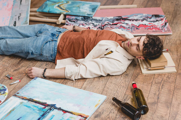 exhausted artist lying on wooden floor, surrounded with paintings, draw utensils and empty bottles