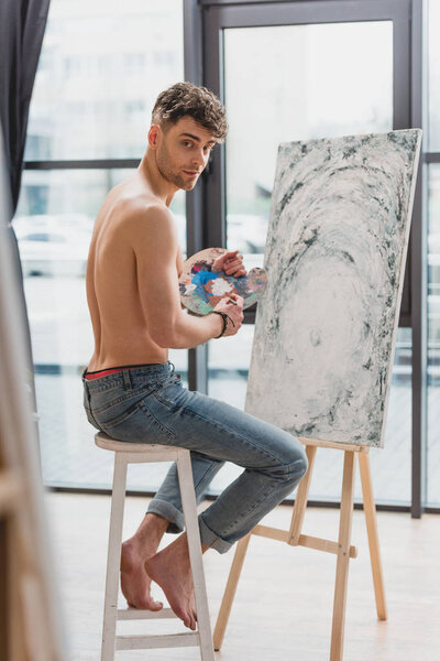 selective focus of good-looking half-naked artist looking at camera while sitting on high chair near easel with painting