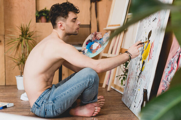 selective focus of thoughtful half-naked artist sitting on floor and painting on canvas 