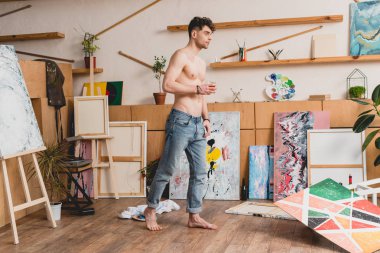 handsome half-naked artist in blue jeans standing in spacious light painting studio clipart