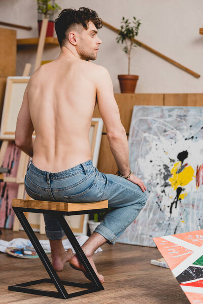 back view of half-naked artist in blue jeans sitting on chair in painting studio
