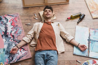 exhausted artist lying on floor in painting studio, surrounded by paintings, empty bottles and draw utensils clipart