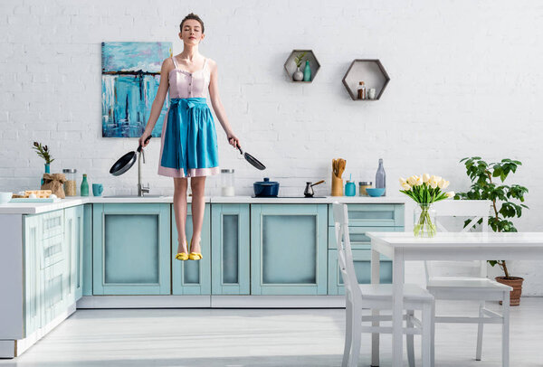 beautiful young woman in apron levitating in air with closed eyes and pans in hand in kitchen