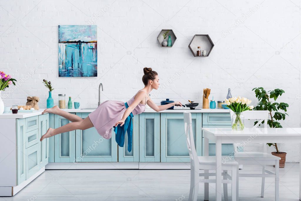 beautiful barefoot girl in elegant dress and apron flying in air with pan in kitchen