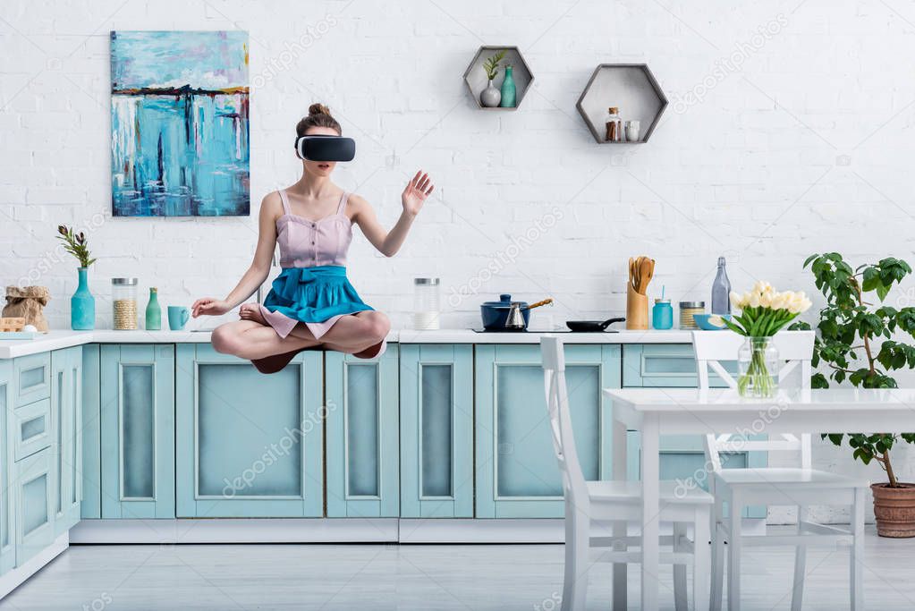 young woman levitating in air in virtual reality headset and gesturing in kitchen