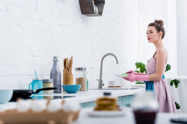 selective focus of young woman in rubber gloves washing dishes in kitchen clipart