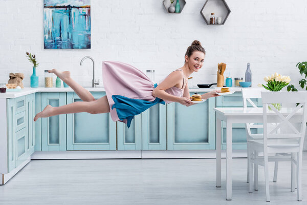 young smiling barefoot woman levitating in air while serving tasty pancakes